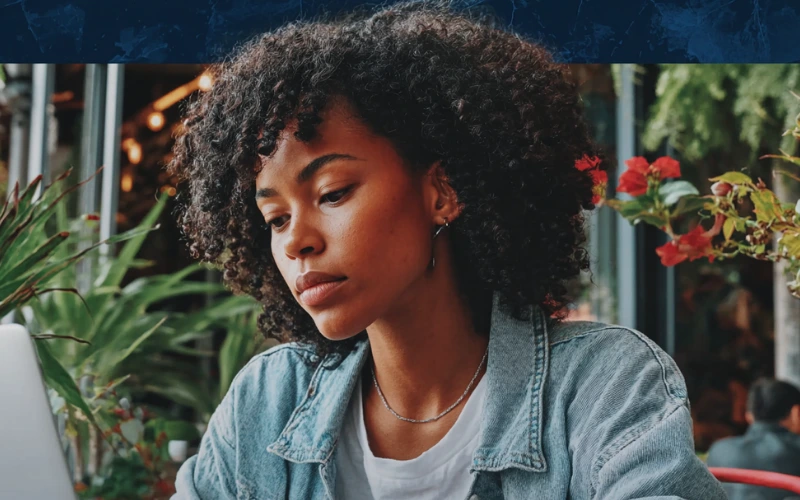 Young woman with natural curly hair in a denim jacket concentrates on a laptop in a bright, plant-filled café or workspace.